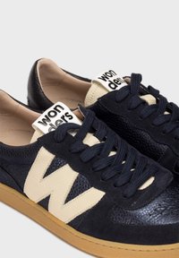 Zapatillas de deporte azul marino con corte de cuero texturizado, detalles en beige y cordones redondos. Presentan un gran logo beige en forma de "W" en el lateral y una etiqueta.