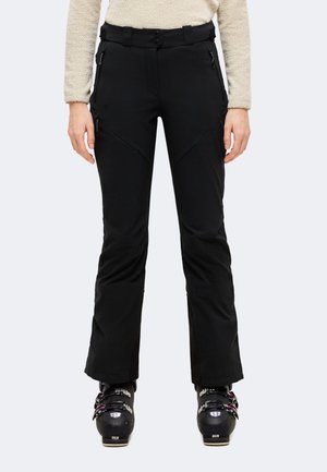 Personne portant un pantalon de ski noir avec des poches zippées et des chaussures de ski, debout devant un fond uni.