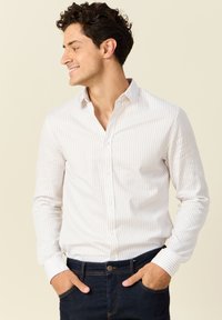 Weißes Langarmhemd mit schmalen vertikalen Streifen in hellem Beige. Verfügt über einen klassischen Kragen und eine Knopfleiste. Dunkle Jeans aus Denim.