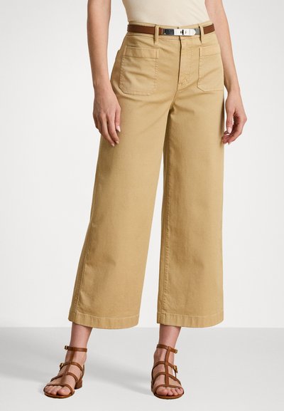 Pantalons larges en khaki en coton, présentant deux poches avant et une fermeture à bouton visible. Associés à des sandales à lanières marron.