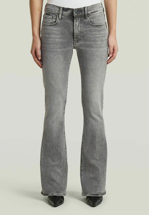 Personne portant un jean bootcut gris et des chaussures noires à bouts pointus sur un fond uni.