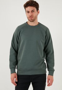 Tmavozelená mikina z mäkkej tkaniny, s okrúhlym výstrihom, dlhými raglánovými rukávmi a malým logom na hrudi.