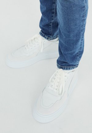 Balti sportbačiai su lygaus odos ir zomšos akcentais, apvaliu nosiu, storu baltu padu ir plokščiomis kryžminėmis raištelėmis; dėvimi su mėlynomis džinsomis.