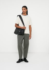 Sort læder skuldertaske med flere sømme, en rummelig design, båret over en neutral t-shirt og parret med løse grønne bukser og sorte sko.