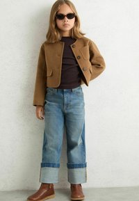 Hnědá vlněná bunda se třemi knoflíky, černý top, světle modré džíny s ohrnutými manžetami a hnědé kotníkové boty. Dítě nosí černé sluneční brýle.