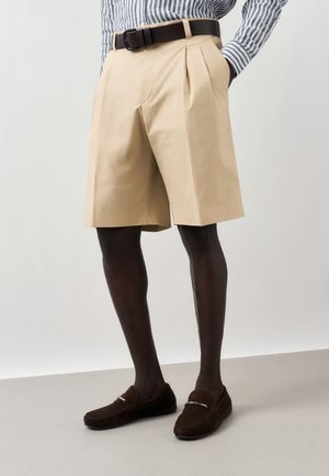 Bézs pliszírozott rövidnadrág sötétbarna mokaszínnal, fekete övvel és kék-fehér csíkos hosszú ujjú inggel párosítva.