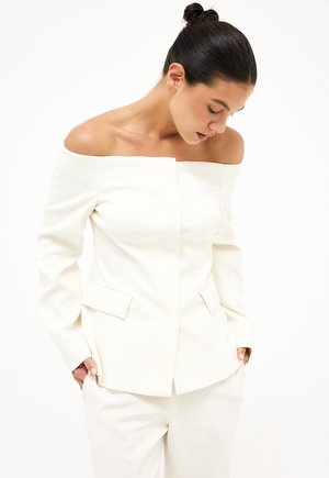 Vrouw met donker haar in een knot, gekleed in een witte blazer zonder schouders en bijpassende witte broek, kijkt omlaag met handen in de zakken.