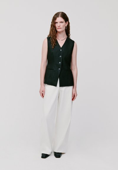 Schwarze ärmellose Weste mit Knöpfen, kombiniert mit weißen weit geschnittenen Hosen und schwarzen Schuhen. Glatte Stoffstruktur, minimalistisches Design.