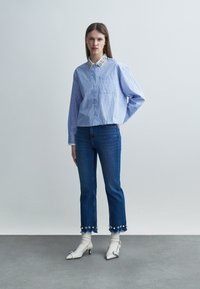 Mujer con camisa azul a rayas con cuello floral, jeans recortados con ribete floral, calcetines blancos y zapatos blancos de tacón bajo, de pie en el interior.