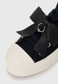 Črne platnene superge z belo gumijasto podlogo, ki vsebuje velik črn usnjen lok, kovinske obroče in temne vezalke. Zgornji del z teksturiranim detajlom.