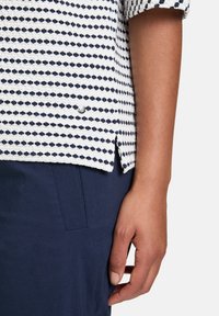 Camicia bianca con motivo geometrico blu navy, in cotone, maniche piegate e spacchi laterali. Abbinata a pantaloni blu navy con dettagli a bottoni.