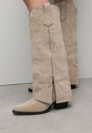 Beige suède kniehoge laarzen met een puntige neus, zichtbare stiksels en een reliëf spiraalpatroon. Voorzien van een zijrits en blokhak.