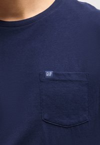 Primer plano de una persona que lleva una camiseta azul marino con cuello redondo y un bolsillo en el pecho con una pequeña etiqueta con el logo de GAP.