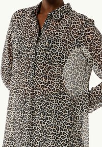 Blouse à imprimé léopard avec col boutonné, manches longues, et tissu léger transparent. Palette de couleurs marron foncé et beige.