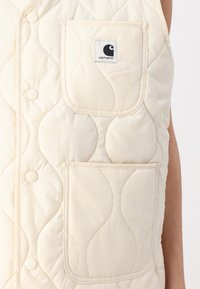 Quiltet kremvest med trykknapper og to frontlommer. Har et Carhartt-logo i en firkantet lap på brystlommen. Strukturerte overflate.