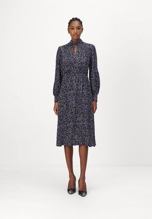 Musta ja violetti kuvioitu mekko, jossa on pitkät puff-hihat, korkea kaulus ja avainaukko-yksityiskohta. Hieman teksturoitu kangas laskeutuu juuri polven alapuolelle.