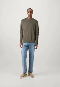 Mann som står iført en olivengrønn genser, lyseblå jeans og beige slip-on sko mot en enkel lys bakgrunn.