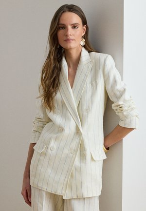 Femme aux longs cheveux bruns portant un blazer croisé à rayures crème et un pantalon assorti, appuyée contre un mur.