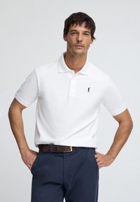 Wit poloshirt met kraag en drie knopen, met een klein marineblauw logo. Gecombineerd met donkerblauwe broek en een bruine riem.