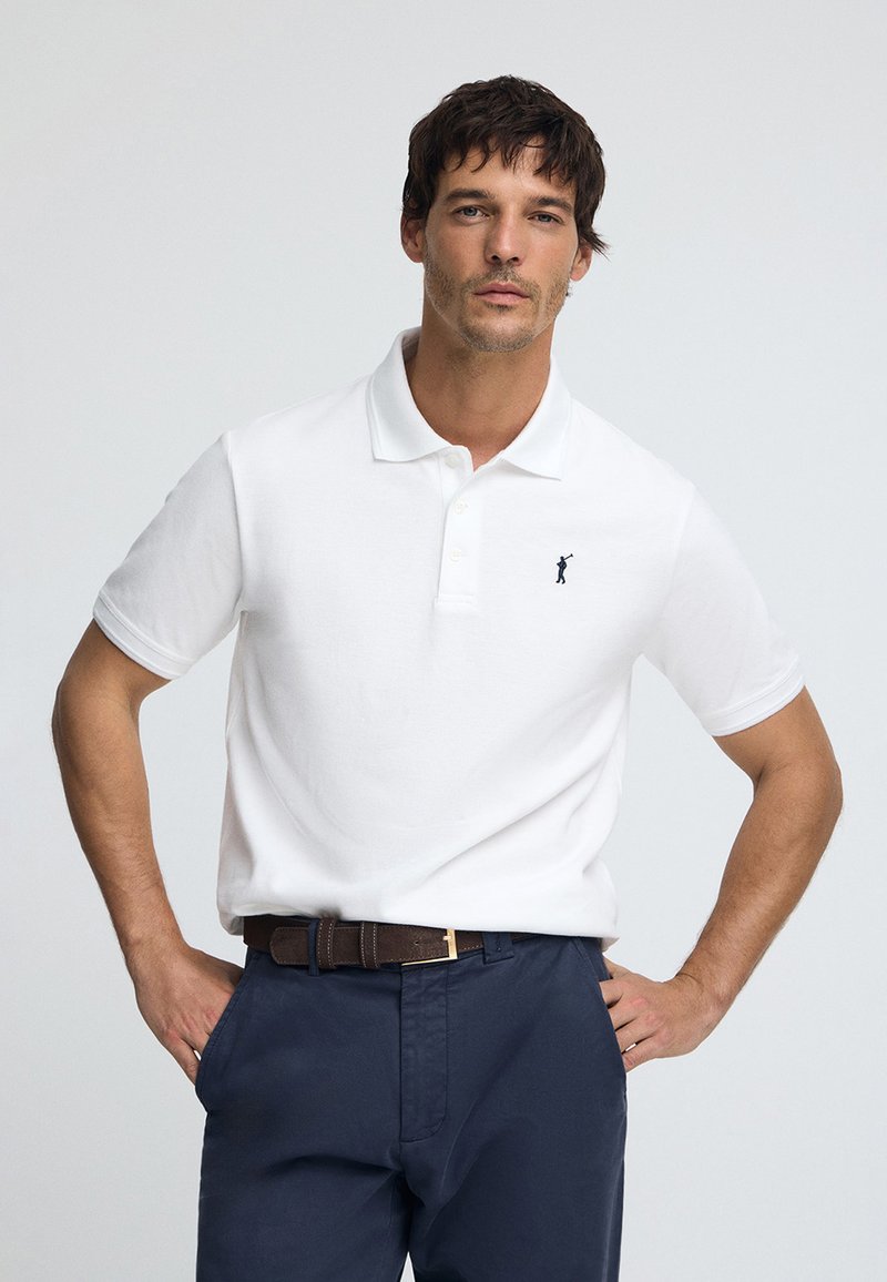 Wit poloshirt met kraag en drie knopen, met een klein marineblauw logo. Gecombineerd met donkerblauwe broek en een bruine riem.