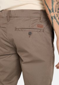 Ruskeat kangashousut, joissa on sileä pinta, varustettuna takataskulla, jossa on nappi, ja pienellä merkkilapulla.