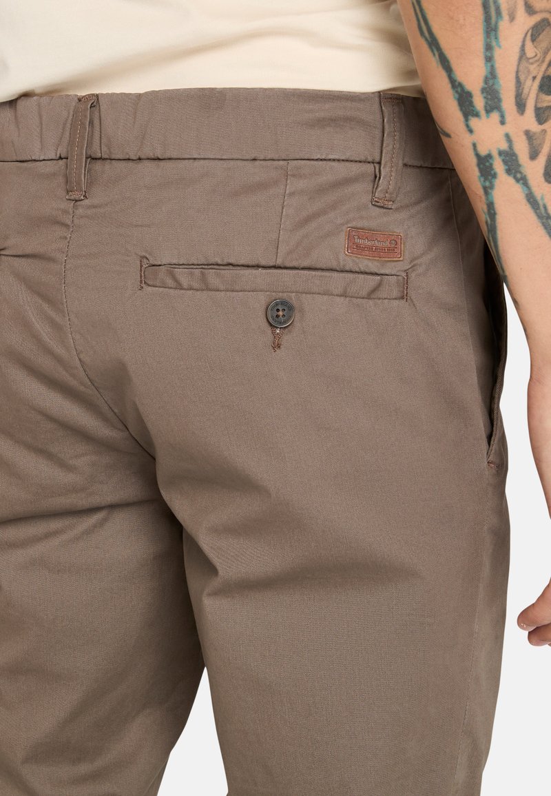 Braune Stoffhosen mit glatter Textur, ausgestattet mit einer Gesäßtasche mit einem Knopf und einem kleinen Markenetikett.