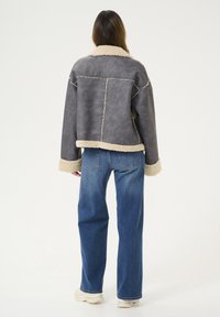 Siva umjetna kožna jakna s bež krznom podstave i manžetama, koja ima kutni dizajn i vidljive šavove, u kombinaciji s plavim trapericama.