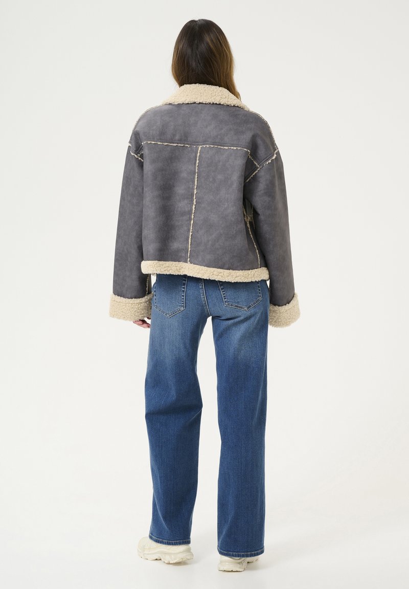 Szara kurtka z sztucznej skóry z beżowym futrem i mankietami, charakteryzująca się luźnym krojem oraz widocznymi detalami szwów, w połączeniu z niebieskimi dżinsami.