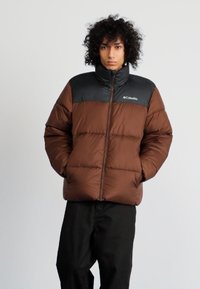 Chaqueta acolchada marrón con sección superior negra, cuenta con cierre de cremallera, diseño acolchado horizontal y logotipo en la parte izquierda del pecho.