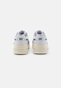 Bílé a navy tenisky s hladkým koženým svrškem, polstrovaným jazykem a límcem, a béžovou texturovanou gumovou podrážkou. Logo ASICS na patě.
