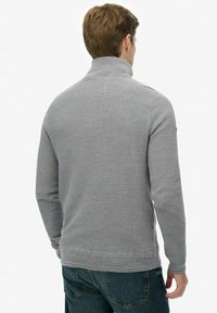 Grijze gebreide trui met hoge kraag, gestructureerd patroon en ribbels bij de manchetten, gecombineerd met blauwe jeans. Achteraanzicht dat het ontwerp en de details toont.