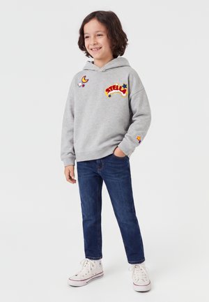 Enfant souriant, portant un sweat à capuche gris avec des patches colorés, jean bleu, baskets blanches montantes, debout avec une main dans la poche sur fond blanc.