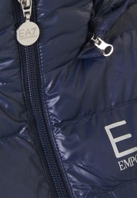 Chaqueta de plumas navy con acabado brillante, costuras en contraste, cremallera completa y logo EA7 Emporio Armani en plata en la parte frontal.