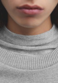 Pull gris en tricot avec un col haut et superposé. Le tissu semble doux avec une texture fine, présentant un motif côtelé régulier en bas.