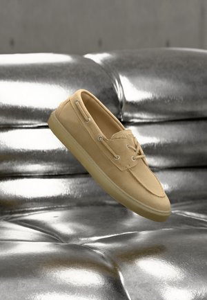 Beiger Wildleder-Bootsschuh mit Schnürdetails, der über einer glänzenden, metallisch silbernen, gesteppten Lederoberfläche schwebt.