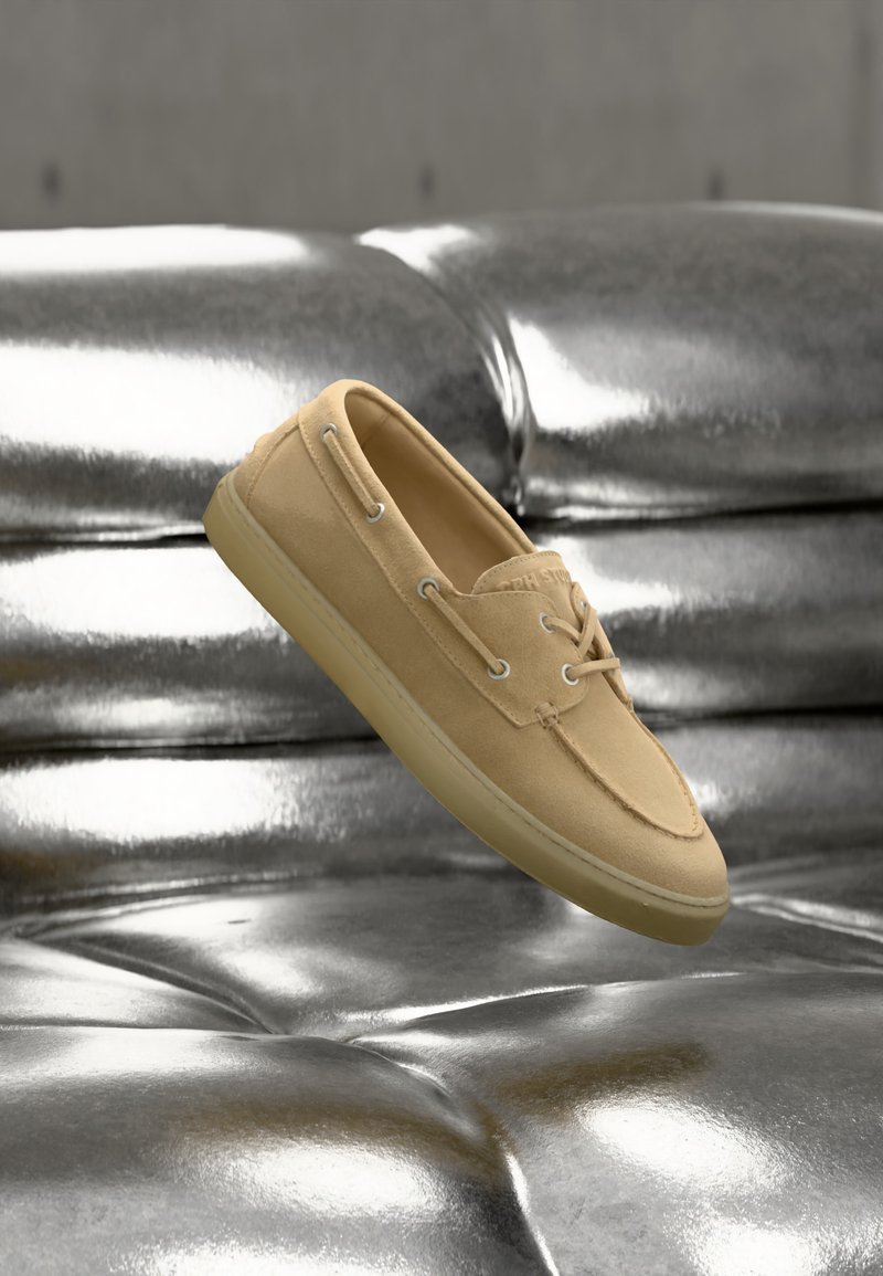 Μπεζ μοκασίνι από καστόρι με λεπτομέρειες κορδονιών, που αιωρείται πάνω από μια γυαλιστερή μεταλλική ασημένια δερμάτινη επιφάνεια με ταφτ.