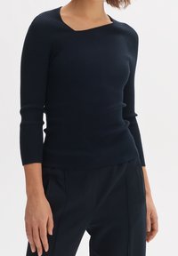 Kvinna i en åtsittande, ribbad mörkblå tröja med trekvartsärmar och mörkblå skräddarsydda byxor som står mot en vit bakgrund.