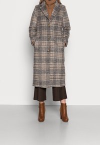 Kostkovaný vlněný kabát v šedé, béžové a hnědé barvě, s klopovým límcem, zapínáním na knoflíky a postranními kapsami, v kombinaci s hnědými botami na block heels.