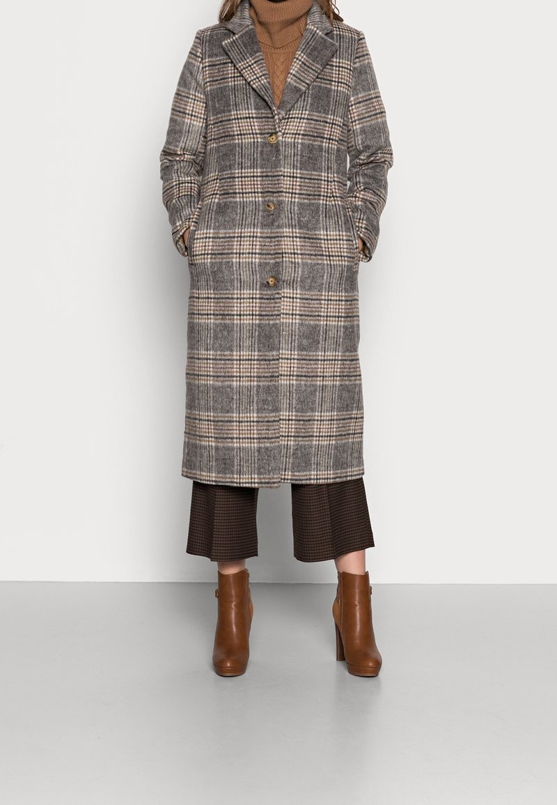 Kostkovaný vlněný kabát v šedé, béžové a hnědé barvě, s klopovým límcem, zapínáním na knoflíky a postranními kapsami, v kombinaci s hnědými botami na block heels.
