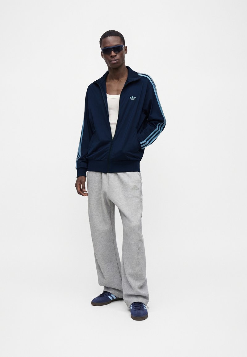 Homme portant une veste de survêtement Adidas bleu foncé, un pantalon de survêtement Adidas gris clair, des baskets Adidas bleues, un débardeur blanc et des lunettes de soleil foncées, debout avec une main dans la poche.
