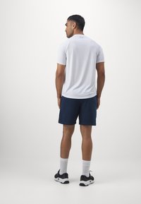 Mand stående med ryggen til iført en hvid kortærmet skjorte, marineblå shorts, hvide strømper og sorte atletikskøjter på en ensfarvet baggrund.