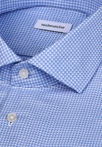 Chemise bleu clair à motif zigzag fin, boutons blancs, et une étiquette blanche portant la mention "seidensticker" à l'intérieur du col.