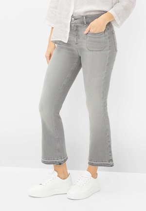 Frau trägt hellgraue Cropped-Flared-Jeans, weiße Sneaker und ein weißes Hemd mit Knöpfen, Hand in der Tasche.