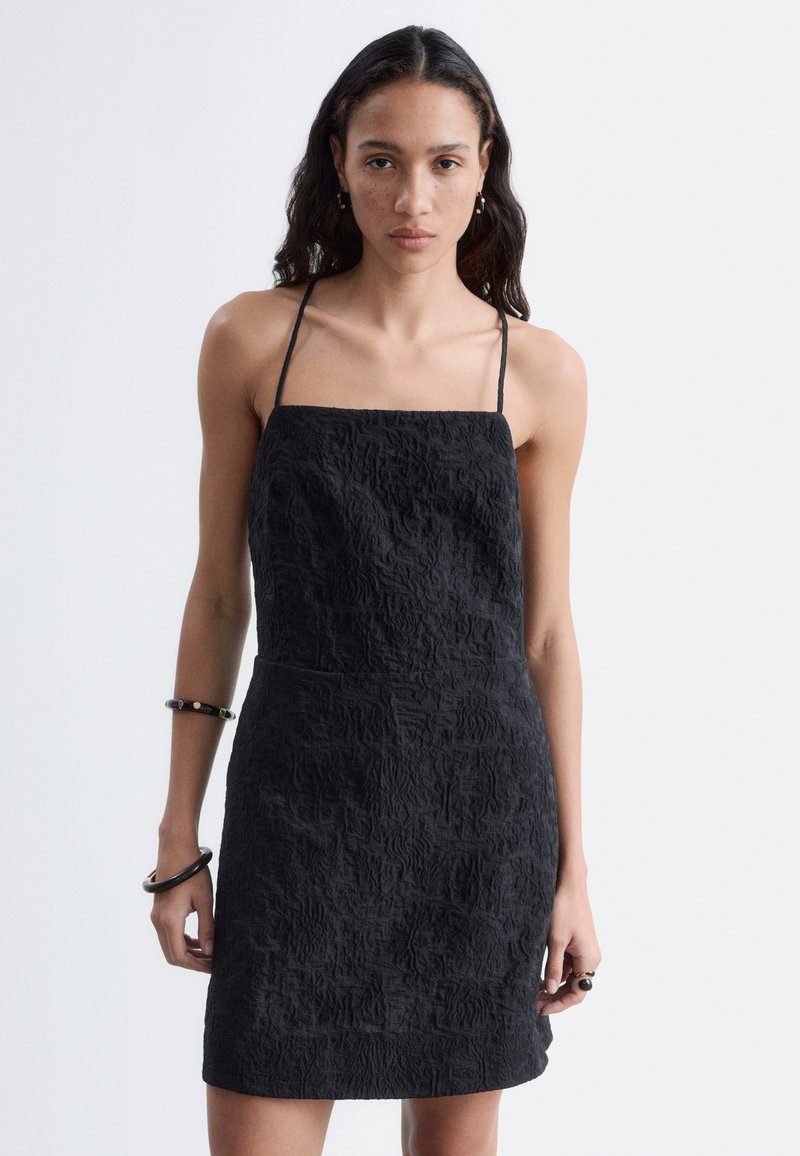 Femme aux longs cheveux foncés portant une robe noire texturée sans manches avec de fines bretelles et des bracelets noirs aux deux poignets, avec une expression neutre.