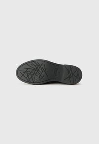 Black rubber shoe sole with textured grip pattern, featuring cross-hatch designs and branding detail. Flat shape with rounded edges.