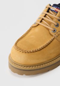 Hellbraune Wildleder-Mokassin-Schuhe mit dicker Sohle, beigen Schnürsenkeln und Metallösen, die detaillierte Nähte und ein markiertes Stofftag zeigen.