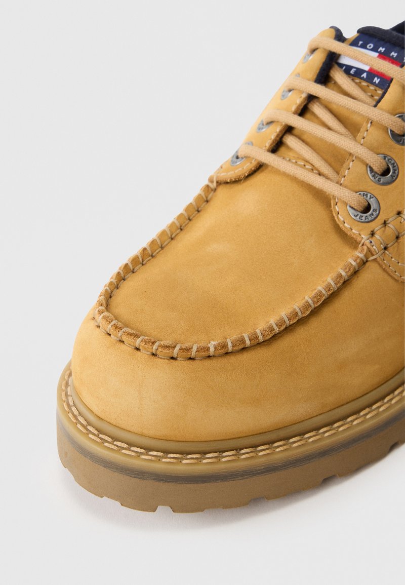 Scarpa moccasin in suede color tan con suola spessa, lacci beige e occhielli in metallo, che mostrano cuciture dettagliate e un'etichetta in tessuto con marchio.