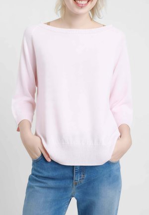 Giovane sorridente con le mani nelle tasche, indossa un maglione rosa chiaro e jeans blu su sfondo semplice.