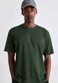 T-shirt en coton vert à manches courtes et encolure ronde. Présente un logo BOSS subtil au centre. Accompagné d'une casquette grise.