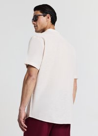 Chemise à manches courtes, de couleur beige clair avec une texture subtile, coupe décontractée et ourlet droit, portée avec un pantalon rouge foncé. Lunettes de soleil présentes.